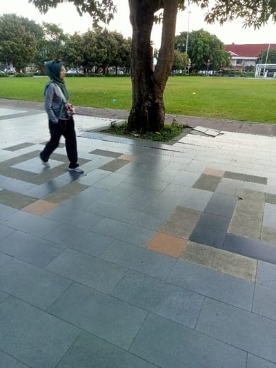 A woman wearing a teal headscarf and a teal bag walks on a tiled path. The path is made of large grey tiles, with a few brown tiles interspersed. The path is surrounded by a lawn area, with a large tree in the middle of the photo. Behind the lawn area, a road with some small buildings on either side can be seen. The scene is located in Pati, Indonesia, at the coordinates -6.754, 111.04.