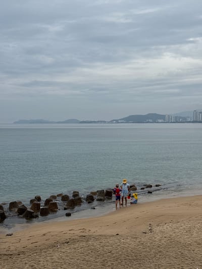 A family of three, two adults and a child, are standing on a sandy beach by the ocean. The adult on the left is wearing a blue and white striped shirt, red shorts, and a yellow sun hat. The adult in the middle is wearing a light blue top, blue shorts, and a yellow sun hat. The child on the right is wearing a yellow sun hat and a blue outfit and is sitting in the sand. They are all facing away from the camera, towards the water, and appear to be engaged in some beach activity. The water is calm and a light blue-grey color, with a line of large, dark, angular rocks breaking the surface near the shore. In the distance, across the water, is a coastline with hills and buildings, including a cluster of tall, modern buildings that suggest an urban area. The sky is overcast with grey clouds. The sand on the beach is light brown and appears somewhat damp near the water's edge, with some footprints visible. This scene is likely located in Nha Trang, Vietnam, given the presence of the coastal city and hills.