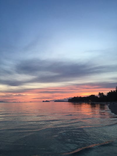 The image shows a tranquil beach scene at sunset in Ban Tai Subdistrict, Thailand. The sky transitions from a soft blue at the top to vibrant hues of orange and pink near the horizon, with wispy clouds scattered throughout. The calm ocean reflects the colorful sky, creating a shimmering effect on the water's surface. In the distance, several small islands are visible, silhouetted against the sunset. The shoreline is lined with dark palm trees and vegetation, and a sandy beach stretches along the edge of the water. A few distant lights can be seen among the trees, suggesting a settlement or resort. The overall atmosphere is peaceful and serene, capturing the beauty of a tropical sunset.