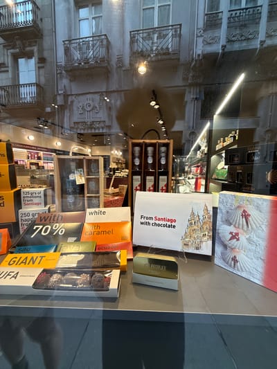 This media file shows a close-up, horizontal view of a shop window displaying various chocolate and honey products. The shop appears to be located in Santiago de Compostela, Spain, as indicated by text on the packaging. The products are arranged on shelves and boxes, with some products featuring imagery of the Santiago de Compostela Cathedral.
Visible text includes "From Santiago with chocolate," "125 grs of emotions from Santiago," "250 grs happ fro," "70%," "white," "with caramel," "ecuador," "THE AMULET TANZANIA 75% DARK," and brand names like "Aialma MEL DE MONTAÑA" and "casal+ coton."
The scene is indoors, with artificial lighting illuminating the products. Reflections on the glass window show the exterior of a building with balconies and architectural details, suggesting an urban setting. The overall mood is inviting, showcasing artisanal food items. The time of day is not discernible, but the lighting suggests it could be evening or a dimly lit interior.