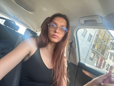 A woman with long, auburn hair and glasses is seated in the passenger seat of a car, looking directly at the camera. The car's interior is visible, including a gray fabric grab handle above the window. Through the window, a building with multiple floors, balconies, and closed red awnings is reflected. The scene appears to be set during the daytime, with bright sunlight indicating clear weather. The woman's expression is neutral. The image is slightly distorted due to the car's curvature and the camera's angle, with the woman's head positioned towards the bottom of the frame.