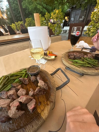 Two wooden platters of food are laid out on a brown tablecloth on an outdoor table. The platters contain sliced steak and green beans. One platter is closer to the foreground and has a small dish of dipping sauce next to it. In the background, a glass of dark liquid with a logo is visible, along with a glass of white wine. The scene appears to be set in a restaurant with greenery visible in the background, suggesting an outdoor dining area. The lighting suggests it is daytime, possibly late afternoon or early evening. A person's hands are visible in the upper right and upper left corners of the image, suggesting people are present and dining. Text visible on the dark liquid bottle reads "PASTA". Other text on a dark structure in the background reads "COFFEE" and "BOLA".