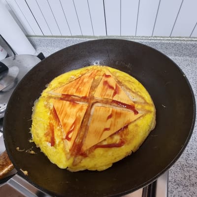 A top-down view shows a circular, golden-brown omelet in a black, slightly worn frying pan. The omelet is topped with four triangular slices of processed cheese, arranged in a way that forms a cross shape. Red ketchup has been drizzled over the cheese and omelet. The omelet appears to be on a stovetop, indicated by a metal grate visible at the bottom of the frame. The background features a grey speckled countertop and white tiled walls with thin, dark grey horizontal lines. The image appears to be taken in a domestic kitchen setting.