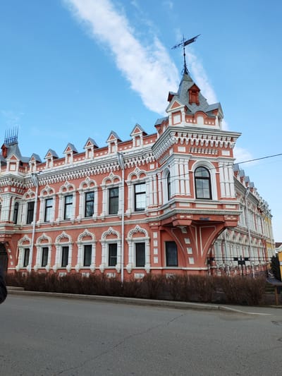 The image depicts the Dacha Bashenina, an ornate pink and white building located in Sarapul, Russia. It is a three-story structure with decorative white trim, arched windows, and a prominent corner turret topped with a weather vane. The building is positioned next to an asphalt road and a low row of bushes. The sky is blue with wispy white clouds. The image is taken from a low angle, emphasizing the building's height and architectural detail. The overall scene suggests a clear, bright day in a quiet urban setting.