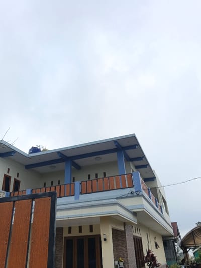 The scene depicts a two-story residential building under an overcast sky, located in Clacuc, Timor-Leste. The building features cream-colored walls with blue accents on the second-floor pillars and balcony railing. The balcony railing also incorporates brown, wood-grain panels. A blue water tank is visible on the flat roof section. The ground floor entrance has dark-framed glass doors, and a section of the ground floor exterior wall is clad with light brown textured tiles. A brown, wood-grain gate, partially open, occupies the left foreground. Utility lines are visible traversing the frame on the right side. Sparse vegetation, including bare tree branches, is present on the left and right sides of the image. No individuals are visible, and no actions or interactions are occurring.