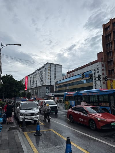 The image depicts a street scene in Yuexiu District, China, under an overcast sky. In the foreground, a wet asphalt road is visible with yellow painted lines and traffic cones. A red taxi and a police van are parked on the side of the road. The police van has "POLICE" written on it in English and Chinese characters, and a police light bar is on its roof. Several people are on the sidewalk, including one person walking with luggage. Further down the street, a blue bus is driving, and various buildings line the road, some with prominent signage in Chinese. The buildings appear to be commercial establishments, with a mix of modern and older architecture. Traffic lights and electrical wires are also visible. The overall impression is a busy urban environment on a cloudy day.