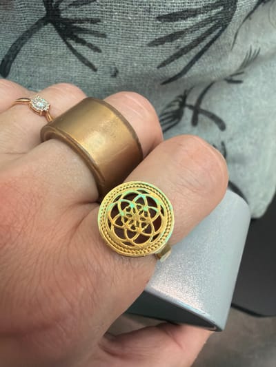 A close-up, overhead shot shows a left hand wearing multiple rings against a textured, gray fabric background with black linear patterns. The index finger is adorned with a wide, brushed-gold band. The middle finger wears a rose gold ring with a square-shaped diamond setting surrounded by smaller stones. The ring finger is decorated with a gold ring featuring an intricate, circular, filigree design with a reddish-brown stone set within. The hand is positioned over a light gray, cylindrical object with a textured surface. The lighting is bright, casting subtle shadows. The image contains no discernible features indicating a specific location in Yekaterinburg, Russia.