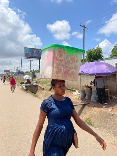 A woman walks down a street in Fegge, Nigeria, carrying a large clear plastic container filled with food on her head. She wears a dark blue dress. In the background, other people walk along the road, and a vendor sits under a purple umbrella selling food. A billboard advertising Guinness is visible in the distance. The street is paved, and there is a gutter on the side. The sky is blue with scattered clouds.