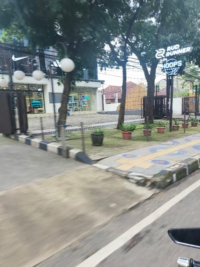The image shows a street in Bandung, Indonesia, located at the coordinates -6.899, 107.621. A black and white checkered curb borders the street, which is paved with grey asphalt. A white line separates the street from a small, grassy area. The grassy area has several potted plants in it, including one in the shape of a basketball. Two large trees are in the background and a large building with a storefront and a storefront with a sign that reads "RUD RUNNER HOOPS". The storefront is open and there is a person standing in the background. The image was captured from a moving vehicle, and the street is blurred. The scene is well lit and the weather is clear.