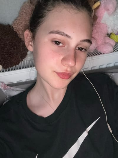 The image shows a close-up, chest-up view of a young female. She has light skin with some freckles, dark brown eyes, and dark brown hair pulled back. Her lips are slightly parted and painted pink. She is wearing a black t-shirt with a white swoosh logo on the left chest. A thin silver necklace is visible around her neck. White earbuds are in her ears, with the wire running down her t-shirt. In the background, there are plush toys, a radiator, and a patterned fabric. The lighting is dim. The scene is indoors. There is no information in the image to determine the location in the city of Panagyurishte, Bulgaria.