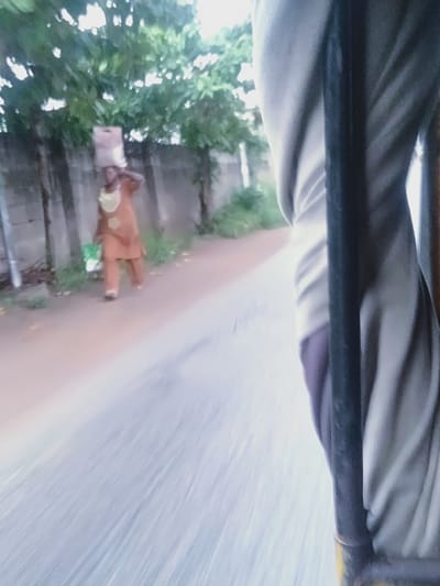 A low-angle, motion-blurred shot captures a person walking on a road in Shangisha, Nigeria. The individual, viewed from behind and slightly to the side, is wearing a traditional orange outfit with a yellow scarf. They are carrying a light-brown package on their head. To the right of the frame, a portion of a person's lower body is visible, wearing gray and purple clothing, suggesting the photo was taken from a moving vehicle. The background shows a rough, grey wall and lush green trees. The road surface is a reddish-brown color with visible motion blur from the vehicle's movement.