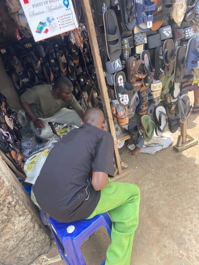 Two men are seen at a street-side stall in Nkpor, Nigeria, selling footwear. The stall is constructed from wood and metal mesh, displaying a variety of sandals and flip-flops. A sign at the top advertises "Point of Sales Services Available," listing options like cash deposit, cash withdrawal, cash transfer, bills payment, and airtime recharge.
The man on the left, wearing a green t-shirt and light-colored pants, is crouched down, attending to items within the stall. He appears to be in the process of organizing or retrieving merchandise. The man on the right, dressed in a black t-shirt and bright green track pants, is seated on a blue plastic stool, facing away from the camera and towards the stall. He is partially obscured by the man on the left.
The ground is paved with dirt and gravel. The background shows more stacked footwear and the rustic construction of the stall. Sunlight illuminates the scene, casting shadows and highlighting the textures of the various materials. The overall impression is of a bustling open-air market environment.