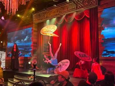 In a dimly lit theatre with a rustic wooden interior, a performance is underway. The stage is adorned with rich red velvet curtains and illuminated by warm stage lights, casting a dramatic glow. Above the stage, a sign reads "Voodoo Child" in ornate script.
The performance features several individuals. Center stage, a woman in a sparkling blue costume is performing an impressive acrobatic pose, balancing on a table with her legs extended upwards. She is surrounded by decorative umbrellas, one held aloft and another open at her side. To her left, another woman in a black dress and veil stands poised, also holding an umbrella. On the right side of the stage, two women in elegant red gowns are seated, one holding a large, decorated umbrella. The backdrop of the stage displays a dynamic digital screen showing what appears to be a stormy or cloudy sky.
The atmosphere suggests an evening performance, possibly a cabaret or a theatrical show, given the costumes and stage setting. The lighting, the ornate details, and the performers' costumes create a sense of theatricality and intrigue. The audience is partially visible in the foreground, suggesting a live event.