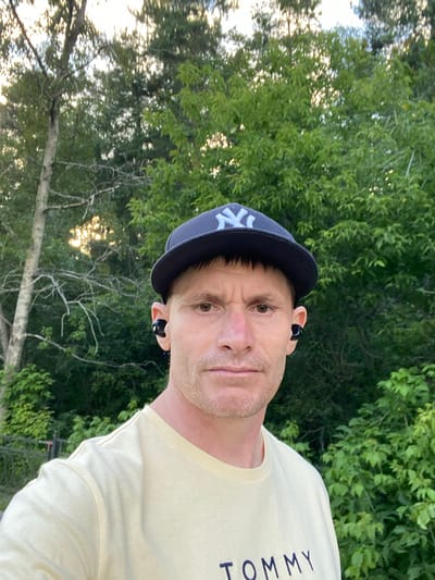 A close-up, selfie-style shot shows a fair-skinned man wearing a light yellow t-shirt with "TOMMY" written on it in dark blue letters, a dark blue baseball cap with a white "NY" logo, and black wireless earbuds. He has short, dark brown hair and is looking directly at the camera with a neutral expression. The background consists of lush green trees and foliage, suggesting an outdoor, possibly wooded, setting. Sunlight filters through the trees, creating a slightly dappled effect. There are no visible landmarks or architectural elements that would indicate a specific location within Yekaterinburg, Russia. The image appears to be taken during daylight hours.