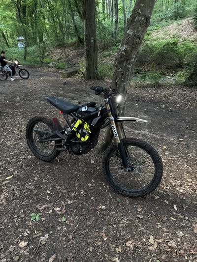 A dirt bike is parked on a dirt path in a forest. The bike is black and yellow, with a single headlight. The path is covered in leaves and dirt, and there are trees on either side. In the background, a person is riding another dirt bike. The scene appears to be a recreational area, possibly in the outskirts of Kichevo, Bulgaria.