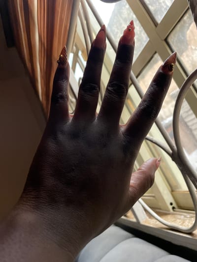 A hand with long, elaborately decorated acrylic nails is held up against a window with metal bars. The nails are painted with a brown and black tiger stripe pattern and adorned with small gold beads and what appear to be small gold charms. The hand itself is dark-skinned. Behind the hand, the window shows a blurred view of what appears to be buildings or structures outside, suggesting an urban or semi-urban environment. The lighting in the image is dim, with light filtering through the window from an exterior source. The curtains behind the hand are a warm, muted brown color. This image was taken inside a residence in Keffi, Nigeria.