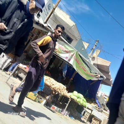 The image, captured in Nuseirat, Palestinian Territory, depicts a street scene with several people in motion. In the foreground, two individuals are partially visible, appearing to be walking past the camera. The focus is on a man in a dark hooded sweatshirt with yellow sleeves and dark pants, walking towards the right side of the frame. His right hand is in his pocket.

Behind him is a market stall covered by a green and white awning with Arabic writing. Underneath the awning, there are fruits and vegetables, including onions and heads of cabbage, displayed on wooden tables. The stall is positioned against a backdrop of buildings, some appearing unfinished. The sky above is clear blue.

Power lines and utility poles are visible against the sky, suggesting the urban setting of Nuseirat. The overall composition of the image suggests a bustling street market in the area.