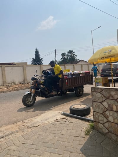 The scene depicts a sunny, clear-day street in Accra, Ghana, characterized by an urban or semi-urban environment. In the mid-ground, a brown motorized cargo tricycle, colloquially known as an 'aboboyaa' or 'pragya', is positioned on the asphalt road, heading left. Its cargo bed is heavily loaded with multiple shrink-wrapped bundles of bottled water or beverages, including packages with red-capped bottles.

Two adult males occupy the tricycle. The driver, wearing a yellow short-sleeved shirt and dark pants, sits at the controls with bare feet visible. A second male, in a dark blue shirt and dark pants, sits in front of the driver, leaning forward, with his face partially obscured.

To the far right, a yellow MTN Mobile Money branded umbrella is erected, shading a small vendor setup. Under this umbrella, a dark-colored SUV (with a visible Ghanaian license plate, e.g., "GH-6435-S") is parked. An individual wearing a light blue t-shirt and dark pants stands near the vehicle, facing away from the camera. A small wooden stand with "MTN Mobile Money" signage is visible under the umbrella.

The foreground features a textured, light-colored stone-clad wall on the right. Immediately next to it on a brick-paved sidewalk (herringbone pattern) lies a black vehicle tire, with a circular metal cover on its hub, and a dark pipe or conduit. In the background, a light-colored concrete block wall runs horizontally, behind which various residential structures and dense foliage are visible. A tall, slender conifer-like tree stands prominently on the left background, with several palm trees further right. Utility poles with overhead lines and a street light pole are also present. The sky is bright blue with a single wispy white cloud. Strong shadows indicate a high sun position.