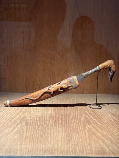 This media file depicts a detailed view of a traditional Vietnamese ceremonial dagger, known as a "dao găm," displayed within a wooden cabinet. The dagger's hilt is uniquely crafted from what appears to be an animal's leg, possibly a deer or goat, with its fur still intact and a curved, dark metal tip. The shaft of the dagger is made of intricately carved wood, featuring flowing, stylized patterns that suggest natural elements like flames or a sinuous creature. Metal accents, possibly silver, adorn the top and bottom of the wooden shaft, providing contrast and detail. The blade of the dagger is visible, partially sheathed in a wooden scabbard. The dagger is suspended by a thin wire hook attached to the wall of the cabinet, casting a distinct shadow. The background is a light-colored, horizontally grained wooden surface, suggesting a well-lit interior space, possibly a museum or private collection. The lighting creates a dramatic effect, highlighting the texture and craftsmanship of the dagger. No people or specific time of day or weather are discernible. The context of Hanoi, Vietnam, implies this artifact may be culturally significant to the region.