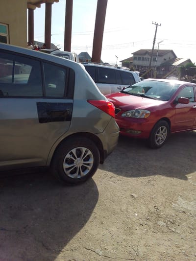 The image shows three parked cars on a dirt and gravel surface. The closest car to the left is a gray hatchback with a dark rectangle covering a portion of the rear passenger door. Next to it is a red sedan. Behind these two is a white SUV. In the background are several buildings and a utility pole. People appear to be gathered on the roof of one of the buildings. The location is Umuode, Nigeria.