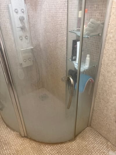 The image shows the interior of a shower stall with a glass door and tiled walls and floor. The shower stall is equipped with a multi-function shower panel. On a glass shelf inside the shower, there are several bottles of toiletries and a light-colored bath glove. The floor of the shower stall appears to be a drain. The overall impression is that of a functional, albeit somewhat dated, bathroom. The provided location of Orekhovka, Russia, is noted for context, but the image itself does not contain any specific geographical indicators to confirm this.