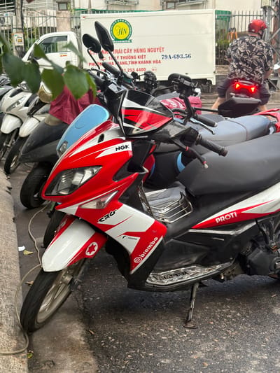 In Nha Trang, Vietnam, a line of parked motorcycles is visible. The focus is on a red and white scooter in the foreground, with the brand "PROTI" visible on its side. Behind it, other scooters are parked, some covered with fabric. A white truck with Vietnamese writing and a circular logo is in the background, suggesting a commercial or delivery context. A person wearing a patterned shirt and a red helmet is seated on a motorcycle further back, facing away from the camera, with its taillights illuminated. The scene appears to be on a street or parking area, with a textured asphalt ground and a concrete curb on the left. Leaves and debris are scattered on the ground.