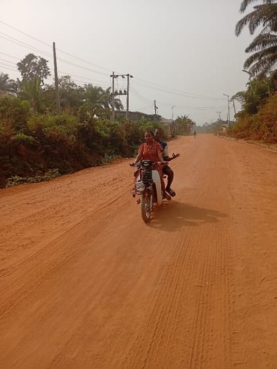 Two people ride a moped on a red dirt road. The road stretches into the distance with vegetation and power lines along the sides. The sky is hazy. The person in the back is waving. Tire tracks are visible in the dirt. A lone figure stands far down the road.