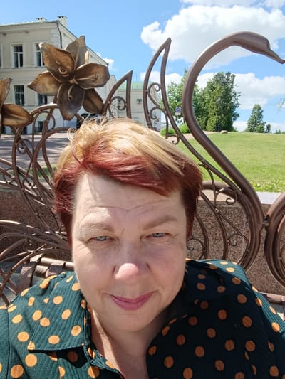 This is a close-up, selfie-style photograph of a woman with short, dyed blonde and red hair. She is seated outdoors, with a decorative metal fence in the foreground and a building and grassy park in the background. The fence features ornate, metallic floral motifs and a sweeping, swan-like shape. The woman is wearing a dark green top with orange polka dots.
The scene appears to be a park or public area, with a building that looks like it could be a historical or institutional structure. The weather is sunny with some fluffy white clouds in a bright blue sky, suggesting it is daytime, likely late morning or afternoon. The woman is looking directly at the camera with a slight smile, conveying a pleasant and relaxed emotion.
The location context places this image in Vitsebsk, Belarus. The overall impression is of a casual outdoor portrait taken on a fair-weather day. There is no visible text in the image.