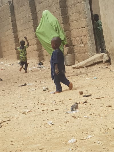 The image depicts an outdoor scene in what appears to be Dutsinma B, Nigeria. Three individuals are visible. One person wears a green garment and stands near a metal container. A second individual, clad in a dark outfit, is in motion, with one foot raised off the ground. A third person, dressed in a green and yellow patterned top, lies prone on the ground. The background is characterized by a light-colored, textured surface, possibly a wall or ground, scattered with debris. In the upper left corner, a person in green lies in a crawlspace. The setting appears to be a public or semi-public space.