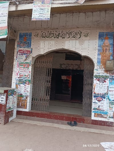 The image displays the street-level entrance of a building in Kaliganj, Bangladesh. An arched doorway is partially open, revealing a dark interior beyond a collapsed grey accordion-style security gate. Above the arch, white tiles feature prominent green Arabic calligraphy, which translates to "There is no god but Allah, Muhammad is the messenger of Allah." This calligraphy is flanked by two tiled illustrations depicting minarets, one on each side. The exterior walls surrounding the entrance are heavily covered with numerous layered paper posters and flyers, primarily in Bengali script, some affixed directly to the tiled surface and others suspended from an overhead overhang. A reddish-brown concrete block, also covered in posters, is situated to the left of the entrance. Two dark-colored sandals are placed on the tiled threshold immediately outside the partially open gate. The ground directly in front of the entrance is unpaved, consisting of dirt or worn concrete. A utility box is mounted on the wall to the right of the entrance. No individuals are visible in the scene. The image is timestamped "04/12/2025 14:07".