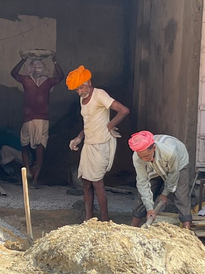 The image depicts three men working at a construction site in Pushkar, India. In the foreground, a man wearing a pink turban and a striped shirt is using a shovel to mix materials in a large pile of what appears to be sand and cement. To his left, another man with an orange turban and a white shirt is standing and looking down, holding a wet cloth in his right hand. Behind them, a third man, his face obscured by a white cloth and wearing a maroon shirt and white dhoti, is carrying a basket filled with materials on his head. The environment appears to be an unfinished building with rough, textured walls and a gravel floor. A wooden plank or pole is visible in the left foreground, propped up against the pile of mixed materials. The lighting suggests it is daytime, possibly mid-morning or afternoon, casting shadows within the structure.