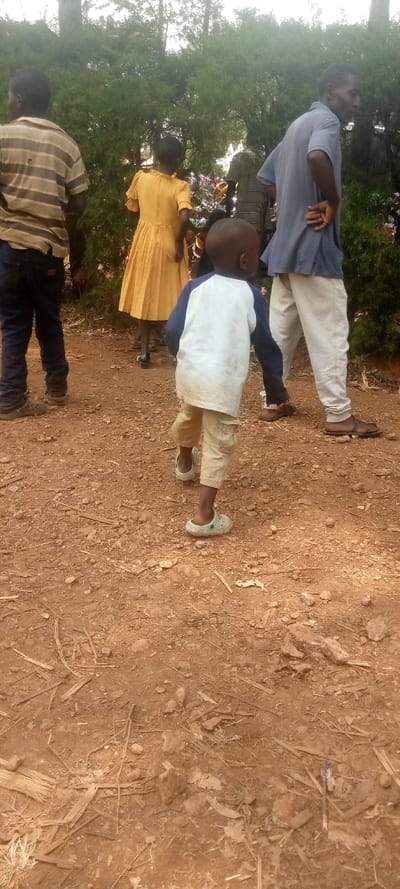 The image shows four people walking on a dirt path. The path is located in a rural area, likely a village. The person in the foreground is a young boy wearing a white shirt and brown pants. He is walking towards the camera, with his right foot forward. He is wearing light-colored sandals. The person behind him is a girl wearing a yellow dress. She is walking away from the camera. Another boy, wearing a striped shirt and blue jeans, is walking toward the camera. In the background, an older man is walking away from the camera. He is wearing a blue shirt and white pants. The man appears to be talking to another person off-camera. The scene is likely captured in Lwandeti, Kenya. The environment is rural and characterized by dirt paths and vegetation. The image appears to be captured during daylight hours.