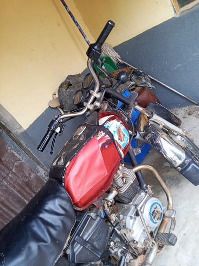 A red and black motorcycle is parked on a concrete surface in Mararaba, Nigeria. The motorcycle is partially covered by a black seat and has a red fuel tank adorned with stickers. Various mechanical parts, including the engine and exhaust, are visible. In the background, a wall painted in alternating bands of yellow and dark gray forms the backdrop. A mop and a blue plastic container are also present in the scene.