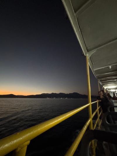 The image depicts a view from the deck of a ferry at dusk, looking out towards a coastline. The sky transitions from a deep blue at the top to a soft orange near the horizon, suggesting the sun has recently set. In the distance, a silhouetted mountain range borders the sea, with faint lights indicating human settlements. The ferry's yellow railing and structural elements are prominently visible in the foreground, suggesting the vantage point is from the upper deck. A few people are standing near the railing, looking out at the view. The water is calm, with gentle ripples reflecting the fading light. The scene suggests a tranquil evening journey. The location is the city of Angra dos Reis, Brazil.