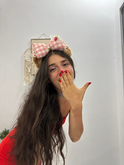 A woman with long, dark hair wears a headband with plush teddy bear ears and a pink gingham bow with a strawberry embellishment. She is dressed in a red top and a sparkly choker necklace. Her right hand is raised to her mouth, covering her lips as if blowing a kiss. Her fingernails are painted bright red. The background is a plain white wall with some decorative items hanging on it, including a macrame wall hanging. A hint of a green plant is visible on the left side of the frame. The lighting suggests an indoor setting. The image does not contain any information indicating its location in Mũi Né, Vietnam.
