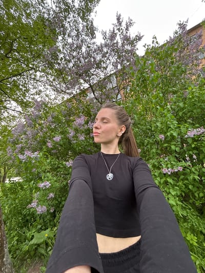 A young woman with light skin and long brown hair is taking a selfie outdoors, likely in a park or garden. She is wearing a long-sleeved black shirt, a necklace with a circular pendant, and an earring on her left ear. She is looking down with her eyes closed. Behind her is dense greenery, including bushes with purple flowers that suggest lilac, and a building is visible in the upper-left corner. The location may be Klin, Russia based on the surrounding vegetation and architecture.