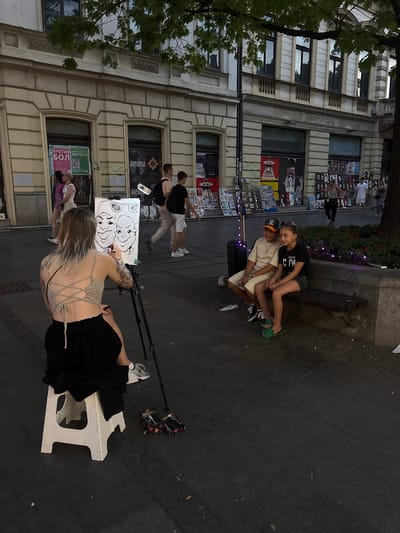 In Belgrade, Serbia, on a paved pedestrian walkway lined with shops and trees, a young woman with blonde hair, wearing a grey lace-up top and a black skirt, is seated on a white stool facing a tripod with a canvas. She is drawing a caricature of two individuals, a man and a woman. The man, seated on a stone bench to the right, is wearing a beige t-shirt and shorts. The woman, seated next to him, is wearing a black t-shirt and shorts. The bench is positioned in front of a raised flower bed. In the background, several people are walking, and various posters are displayed on the shop fronts. Two boxes containing markers are visible on the ground near the artist.