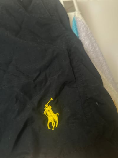The image shows a close-up of a black garment, possibly a shirt or jacket, with a yellow embroidered logo. The logo depicts a horse and rider, characteristic of the Polo Ralph Lauren brand. The fabric appears to be a textured cotton or blend, with visible stitching lines and subtle wrinkles suggesting it has been worn. In the background, out of focus, are hints of other fabrics, including a light gray and a light blue, possibly other clothing items or towels.
The lighting is somewhat dim, suggesting an indoor setting, possibly a closet or a laundry area. There are no discernible location cues beyond the provided context of Gelsenkirchen, Germany. No specific activity or event is taking place; the image appears to be a still shot focusing on the garment's detail. No text is visible in the image. The overall impression is that of a detail shot of clothing.