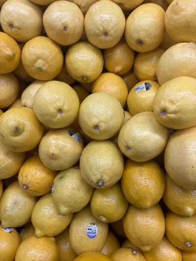 A top-down close-up shot displays a pile of ripe, yellow lemons. Some lemons have small, round indentations where the stem was attached. A few of the lemons have stickers with various markings. The lemons are tightly packed, creating a textured surface. The lighting is bright, highlighting the glossy skin of the fruit. This image was taken at a supermarket in North Jakarta, Indonesia.
