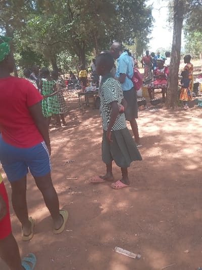 The image shows a group of people standing in a dirt path located in Malava, Kenya (latitude: 0.53, longitude: 34.812). The path is situated under a canopy of trees. There are five people standing in the path, all of whom are facing the camera. Three individuals are wearing red shorts and shirts. Two people are wearing green and white checkered shirts and dark green shorts. Two of the individuals are wearing sandals and flip-flops.  The people in the image appear to be conversing with each other. The ground is brown and dusty, and there are shadows cast from the trees on the ground. There are objects on the ground such as sticks, leaves, and a piece of broken plastic.  There is a group of people in the background sitting on a bench under a tree to the left of the frame.  It is not clear what activity is taking place in this scene.  The image was taken during the day.
