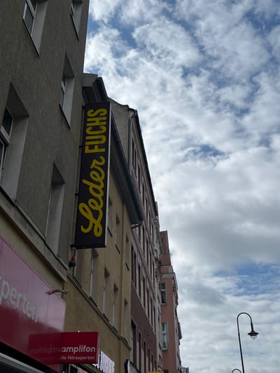 The image shows a vertical sign with yellow text on a dark background that reads "Leder Fuchs." The sign is attached to the side of a building, which appears to be in an urban environment. The building's facade is a neutral gray color with visible windows and trim. In the background, other buildings are visible, some with reddish-brown brickwork. The sky above is filled with clouds, suggesting an overcast or partly cloudy day. The overall scene depicts a street view in a city, likely a commercial area given the presence of the sign and other businesses visible in the background, such as one with a red sign and white text that reads "perten" and another red sign with white text that reads "amplifon Die Hörexperten". The image was taken from a low angle, looking upwards towards the buildings and the sky. The location is in Berlin, Germany.