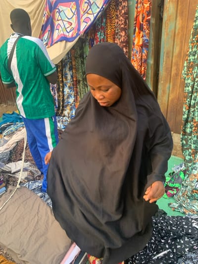 The image depicts an outdoor market scene in Okene, Nigeria, featuring two individuals amidst various fabric displays. In the foreground, a woman of African descent is seen from the chest up, wearing a plain black hijab covering her head and neck, and a loose-fitting black abaya-style garment. Her gaze is directed downwards and slightly to her right, and her right hand is visible near her hip, seemingly adjusting her clothing or reaching for an unseen object below. Behind her, to the left, a man is visible from the back, wearing a green and white striped short-sleeved shirt and blue athletic pants with a vertical green and white stripe.
The background consists of multiple fabric stalls. To the left, several rolls or lengths of colorful, patterned fabrics are draped and displayed, including designs with geometric shapes, animal prints, and floral motifs. A rusty, corrugated metal surface, likely part of a stall structure, is visible to the right of the fabrics. The ground beneath the individuals and stalls is unpaved, appearing as dirt or worn earth, and is scattered with various fabric scraps and debris, consistent with a bustling market environment. The scene is brightly illuminated by natural light.