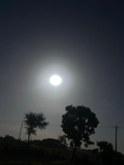 The image displays an outdoor, backlit scene in Rido, Nigeria. The sun, appearing as an overexposed, bright white orb with a prominent halo and a small blue lens flare, dominates the central upper portion of the frame. The sky transitions from bright white around the sun to a dark grey-blue gradient towards the edges. The lower half of the image consists of dark, silhouetted landscape features. These include two distinct trees in the midground: a smaller tree with a sparser canopy on the left, and a larger, denser-canopied tree to the right. A third, smaller, palm-like tree silhouette is visible on the far right. Two utility poles with visible wires are present: one on the left, partially obscured by the smaller tree, and another on the far right. The background features indistinct, mounded terrain or structures, all rendered in dark silhouette. No individuals are visible. The image possesses a soft focus throughout.