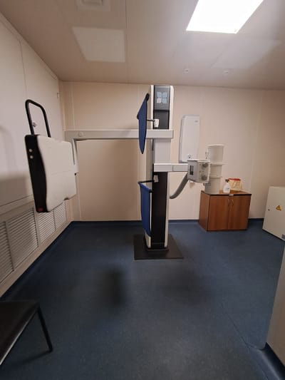 A room, possibly a hospital or clinic in Elektrostal, Russia, contains medical equipment. The equipment includes a large X-ray machine consisting of a diagnostic scanner with blue pads, a white overhead tube structure, and a portable patient stand. A brown cabinet is mounted to the wall above the machinery. The floor appears to be blue vinyl or rubber. A window provides natural light.