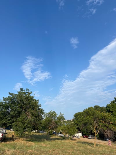 The image depicts a clear blue sky with scattered white clouds, hinting at a pleasant day. The scene is split between the sky and a grassy area with trees. On the right side, a line of trees borders a grassy field. Further back, a dirt road is visible, with several vehicles parked along it, including a white trailer, a red car, a silver SUV, and a darker colored SUV. A person in a light-colored dress with a floral pattern is walking on the dirt road. In the distance, more trees and buildings are faintly visible. The overall impression is that of an outdoor gathering or campsite, possibly in a park or rural area. The lighting suggests it is daytime, likely late morning or afternoon.