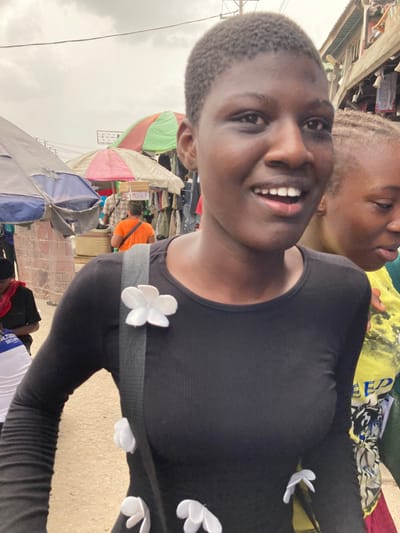 The image depicts a crowded outdoor market in Aba, Nigeria, under an overcast sky. In the foreground, a young Black woman with short, dark, closely-cropped hair smiles with her mouth open, revealing upper teeth. She wears a black, long-sleeved ribbed top embellished with multiple white, flower-shaped fabric appliques. A dark shoulder strap, also bearing a white flower applique, crosses her torso. To her immediate right, a second individual, partially obscured, shows braided hair and a yellow patterned shirt. The midground features market stalls beneath large, colorful umbrellas (blue-grey, red-white, green-brown), displaying hanging merchandise. An individual in an orange shirt is seen from behind, carrying stacked boxes on their head. Overhead power lines traverse the upper frame, and a building with a corrugated metal roof is visible on the far right. The ground appears as an unpaved surface.