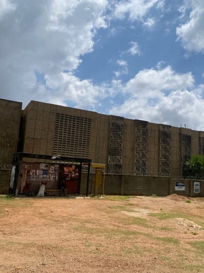 The image captures an outdoor scene in Yelwa, Nigeria, on a partly cloudy day. The focal point is a large, weathered, concrete building with a textured facade and distinct architectural elements, including large rectangular ventilation blocks and rows of circular decorative openings. In the foreground, a dilapidated bus stop shelter is visible, covered in posters and showing signs of wear. A person is seated inside the shelter. To the right of the shelter, a low, barbed-wire-topped wall separates the area from the building. The ground is a mixture of reddish-brown earth and scattered gravel, with some patches of dry grass. The sky is predominantly blue with scattered white cumulus clouds.