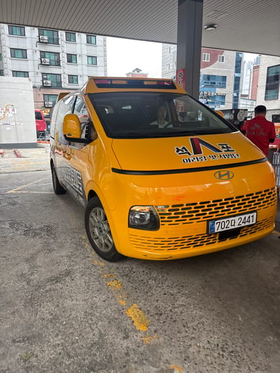 The image shows a yellow commercial vehicle, likely a taxi or delivery van, parked at a gas station. The vehicle has Korean license plates that read "702다 2441". The gas station appears to be an SK Gas station, as indicated by branding on a worker's shirt and signage. The background shows typical urban architecture, including multi-story apartment buildings and other commercial structures. A person is visible in the driver's seat of the vehicle. The scene is consistent with a gas station in an urban environment in South Korea, likely Busan given the presence of SK Gas stations which are prevalent there.