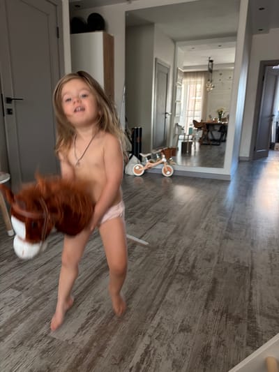 A young girl with blonde hair is straddling a toy rocking horse with a brown mane. She is wearing light pink underwear. Her head is turned towards the camera, and she is smiling. The girl appears to be in a home, with a wooden floor and a large mirror in the background. The mirror reflects a dining area with a table and chairs, and a window with blinds. The scene is lit by natural light, suggesting it is daytime. The overall mood appears playful and carefree. The location is identified as Krasnoyarsk, Russia.