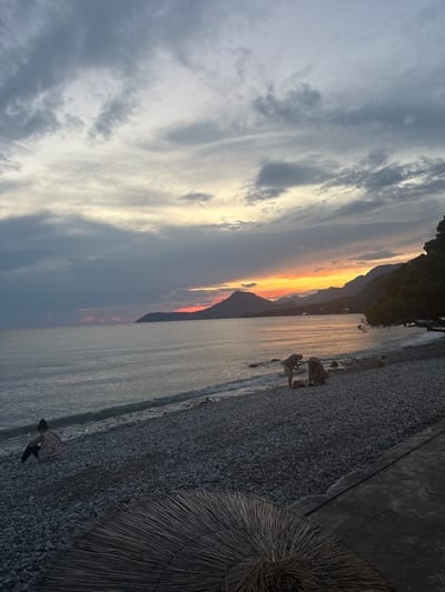 A vertical, wide shot captures a serene sunset scene at a pebble beach in Bar, Montenegro. The sky is a dramatic canvas of swirling gray clouds, dramatically contrasting with vibrant streaks of orange and yellow as the sun dips below the horizon. Mountains, silhouetted against the fiery sky, form a majestic backdrop to the tranquil sea.
On the shore, several people are visible. Two figures are standing near the water's edge, seemingly engaged in an activity. Further down the beach, another person is seated, looking towards the sea. The beach itself is covered in smooth, dark pebbles, with a concrete walkway bordering it. To the right of the frame, a thatched umbrella, a common sight on sun-drenched beaches, adds a rustic touch. The overall atmosphere is peaceful and contemplative, enhanced by the soft light of the setting sun reflecting on the gentle waves. The time of day is clearly dusk, with the fading light casting long shadows and bathing the scene in a warm, diffused glow. The weather appears calm, with a slight ripple on the water indicating a gentle breeze.